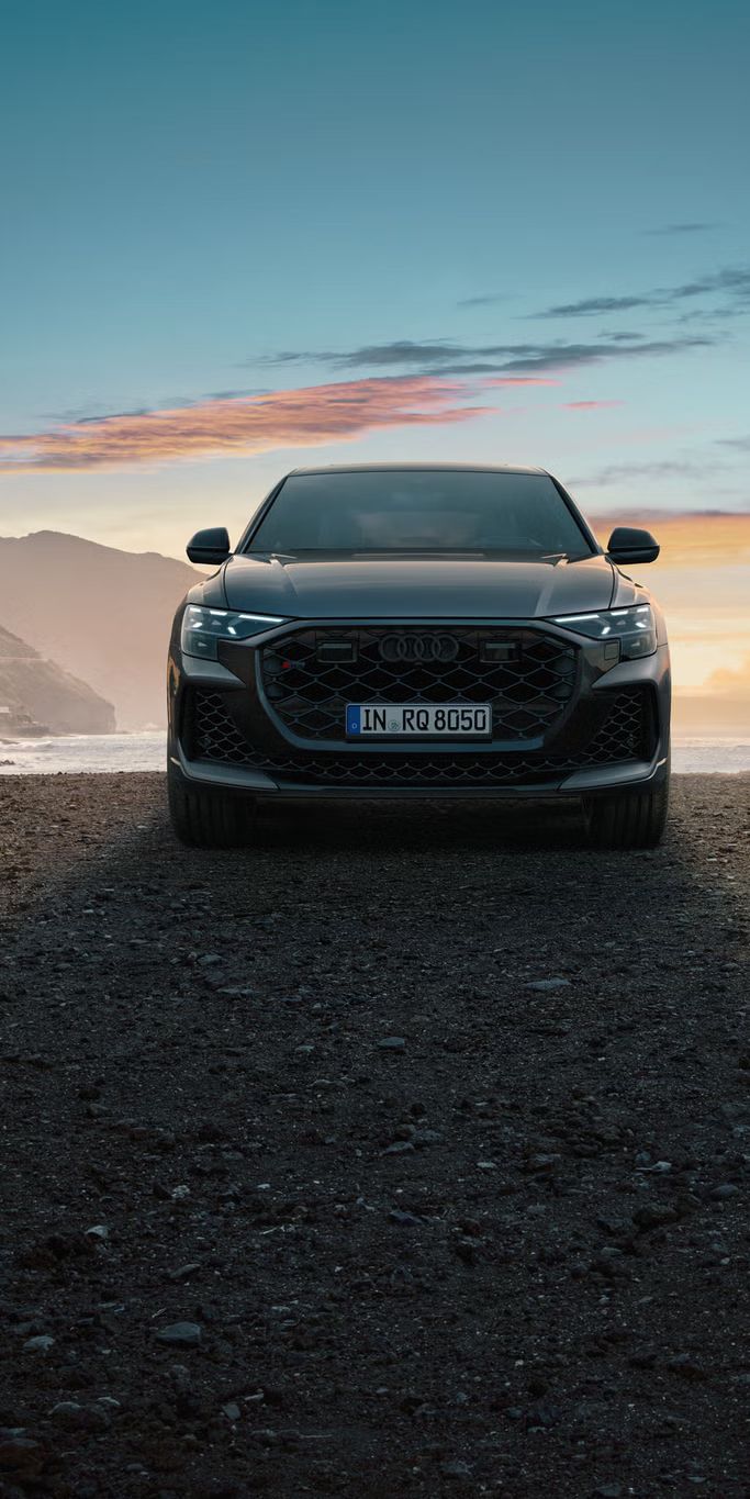 Der RS Q8 SUV von vorn an einem Strand auf Schotter, im Hintergrund das Meer und seitlich hervorragende Berge.