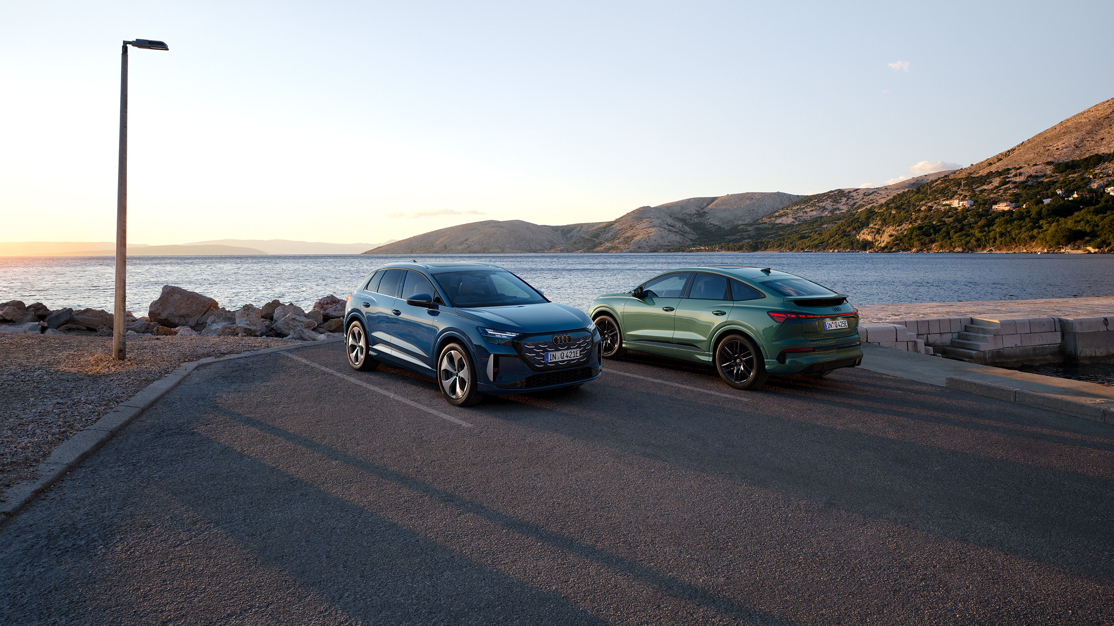 Q4 SUV e-tron quattro performance și Q4 Sportback e-tron quattro performance parcate pe un drum de coastă, cu marea și munții în fundal.