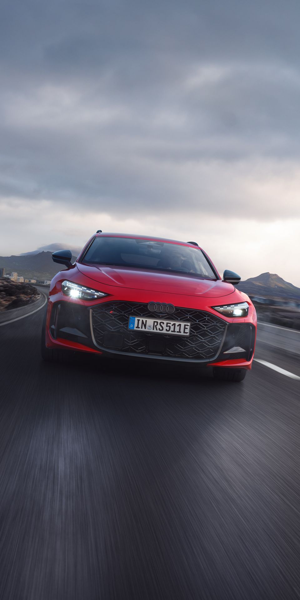 Un Audi RS 5 Avant roșu circulă pe un drum într-un peisaj muntos. Cerul e înnorat.