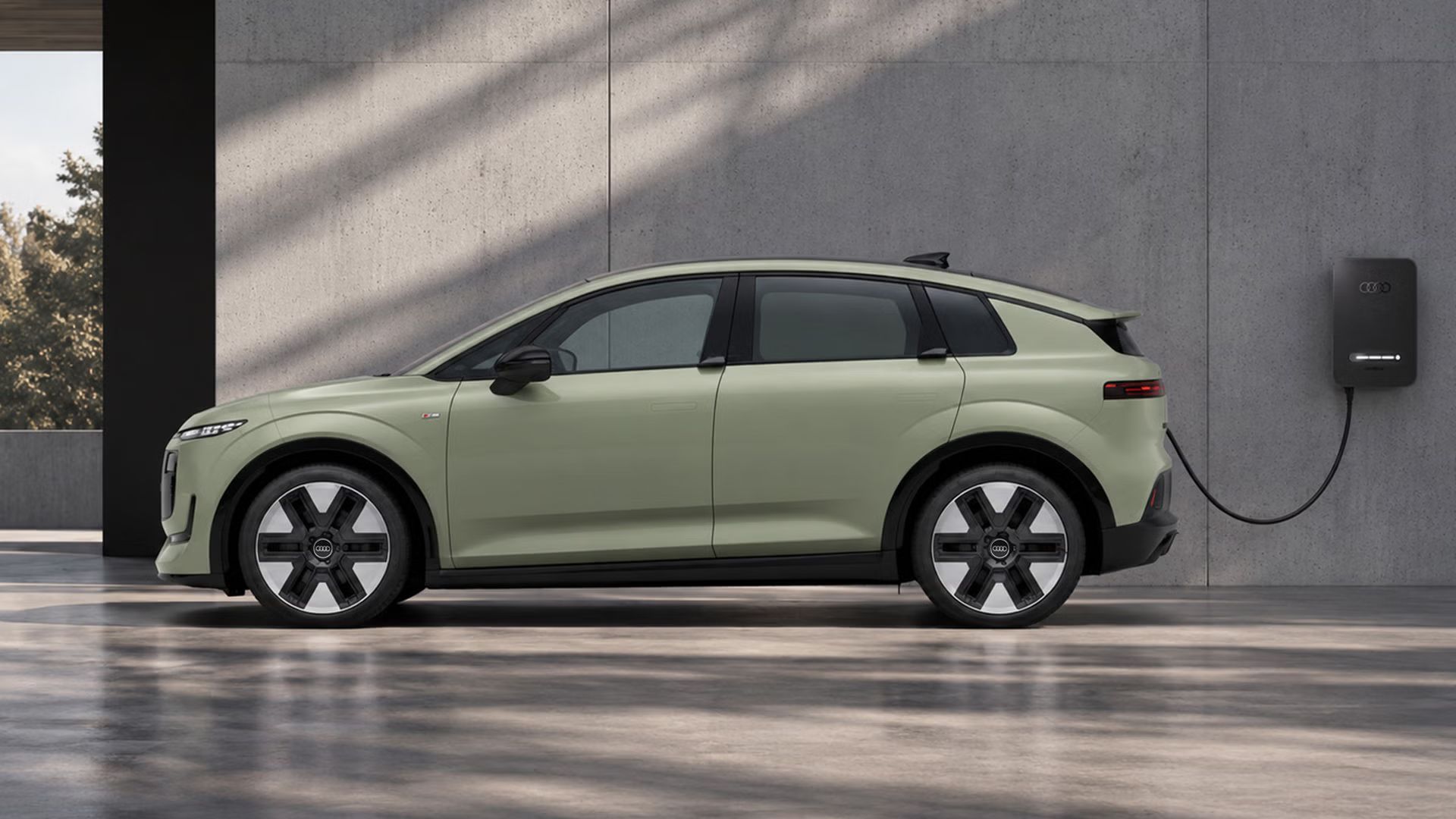 Der Audi A2 e-tron von der Seite parkt vor einem modernen Gebäude in Betonoptik. Das Fahrzeug ist an eine Ladesäule angeschlossen. Licht fällt von oben ein und taucht den glatten Untergrund in eine gescheckte Fläche.