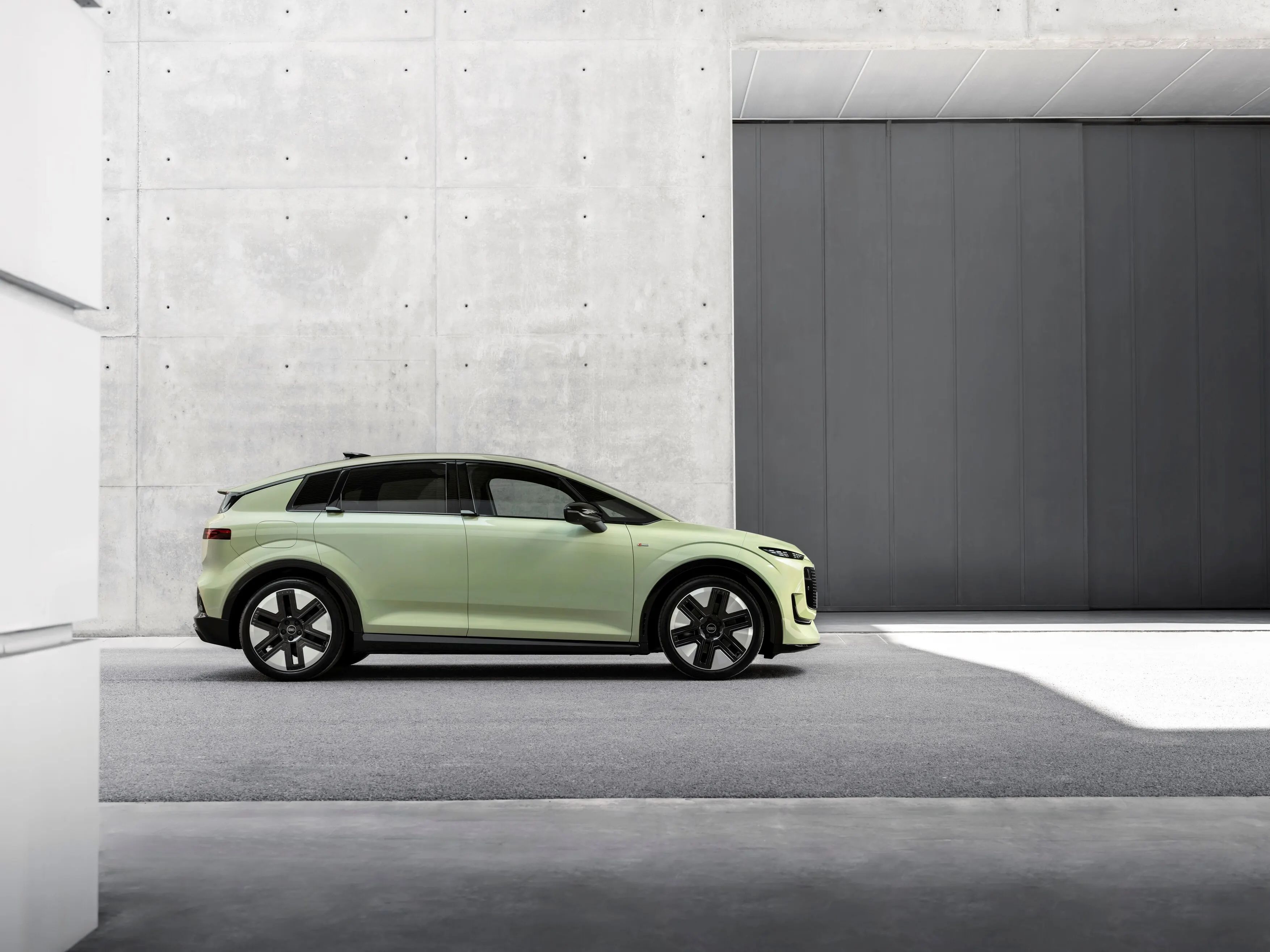 Audi A2 e-tron. Un SUV electric modern, de culoare verde pal, este parcat lateral într-un spațiu urban minimalist, cu pereți din beton gri. Mașina are jante mari, negre, și linii de design fluide și futuriste. În fundal se văd suprafețe netede din beton și o poartă industrială de culoare închisă.
