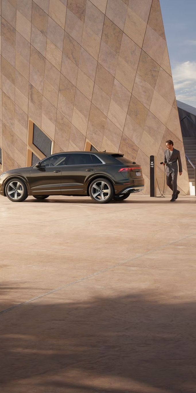 Der Q8 SUV e-hybrid schräg von der Seite parkt vor einem futuristischen Gebäude. Ein modisch gekleideter junger Mann steht hinter dem Fahrzeug und hält den Stecker einer Ladesäule in der Hand.