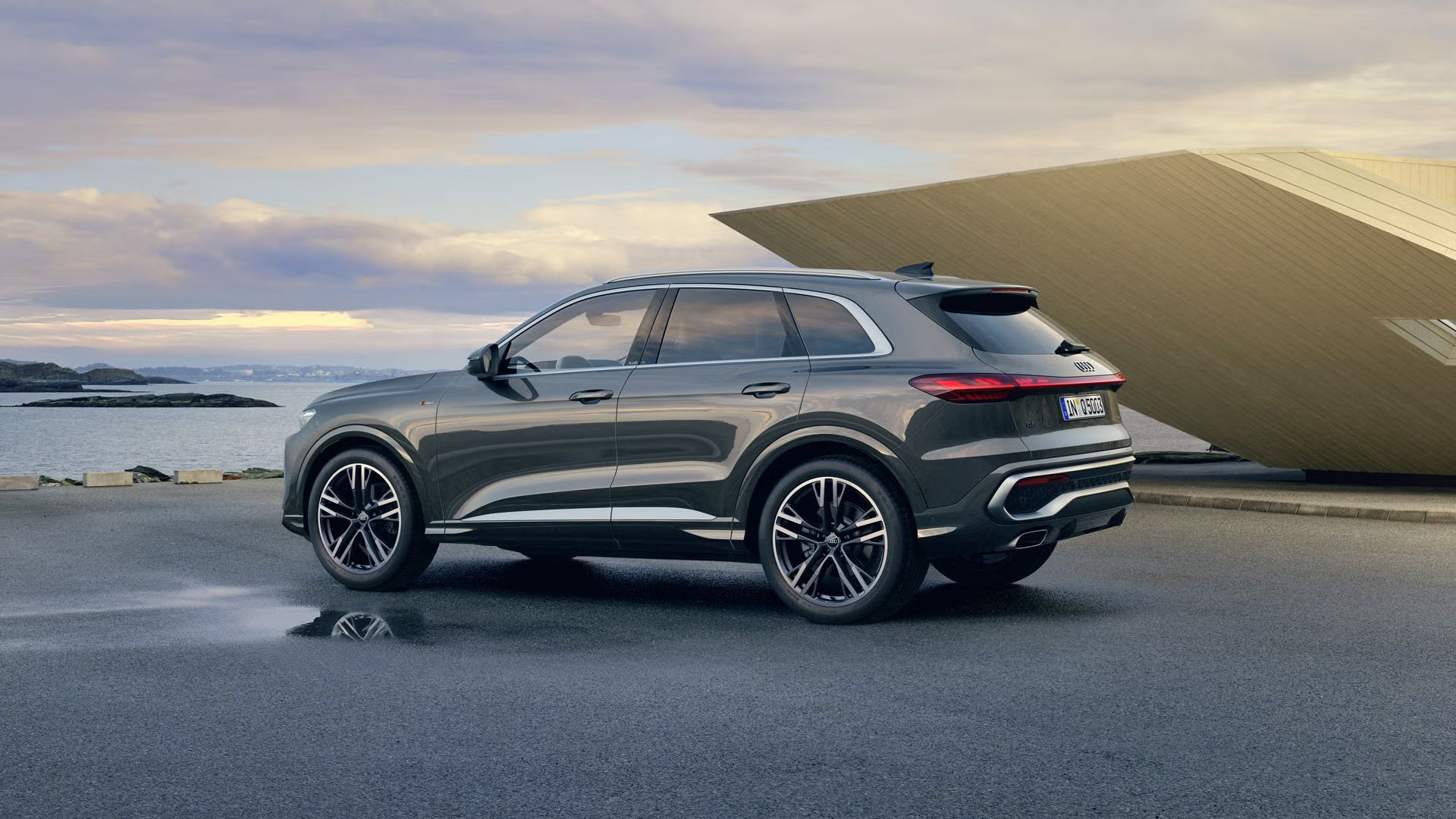 Bočný pohľad na sivé Audi Q5 SUV zaparkované pri futuristickej budove na pobreží.
