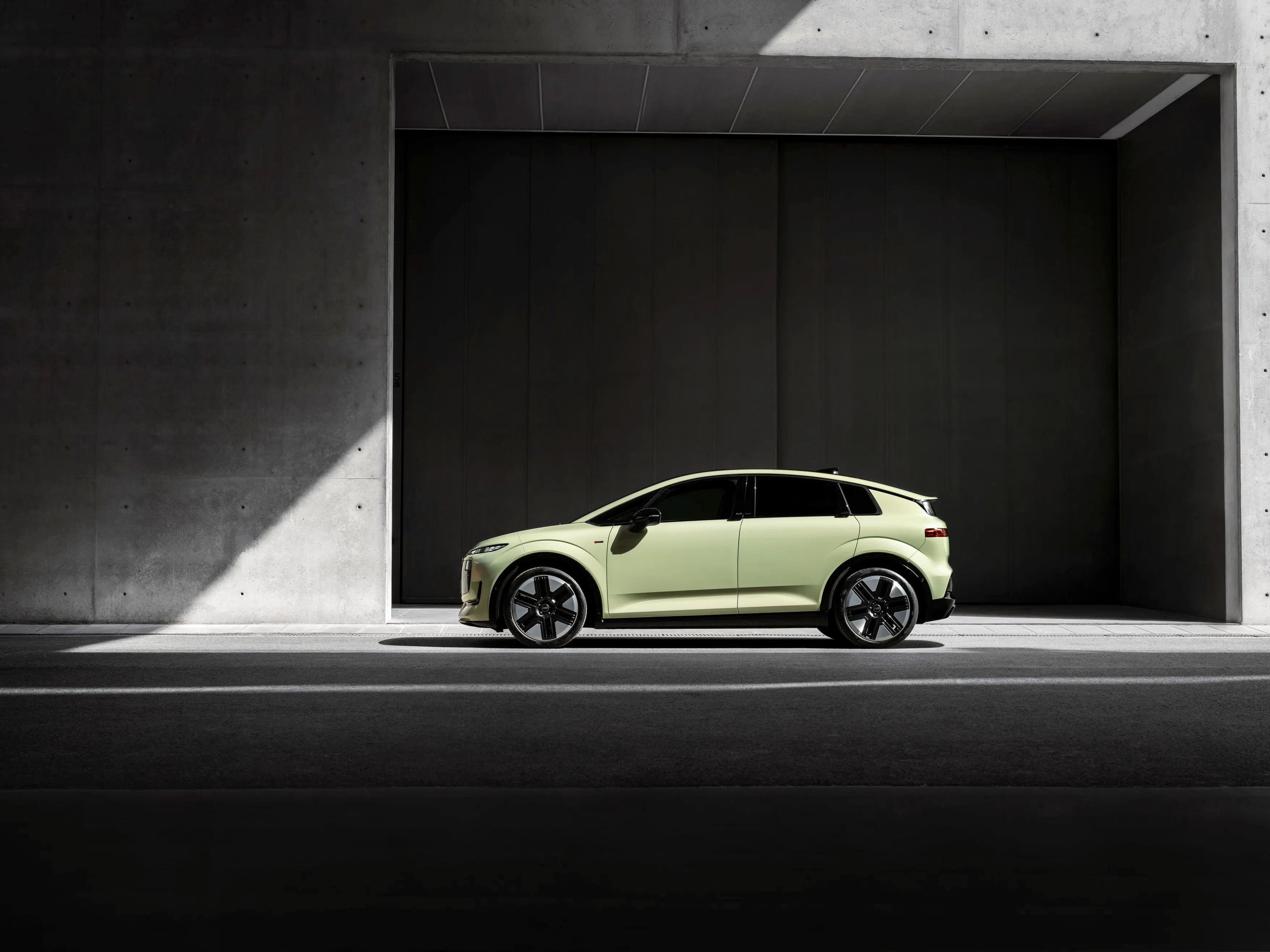  Audi A2 e-tron. Un SUV electric modern, de culoare verde deschis, este parcat într-un cadru urban minimalist din beton gri. Mașina este poziționată în profil, iluminată parțial de o rază de lumină care intră din stânga. Fundalul este compus din pereți înalți de beton și o deschidere întunecată, creând un contrast puternic între lumină și umbră.