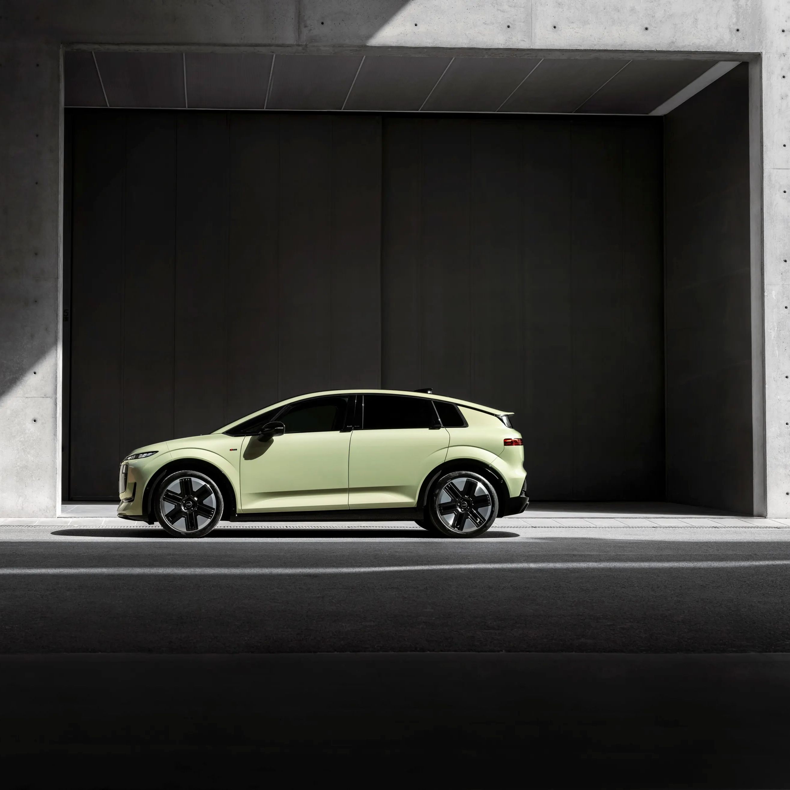  Audi A2 e-tron. Un SUV electric modern, de culoare verde deschis, este parcat într-un cadru urban minimalist din beton gri. Mașina este poziționată în profil, iluminată parțial de o rază de lumină care intră din stânga. Fundalul este compus din pereți înalți de beton și o deschidere întunecată, creând un contrast puternic între lumină și umbră.