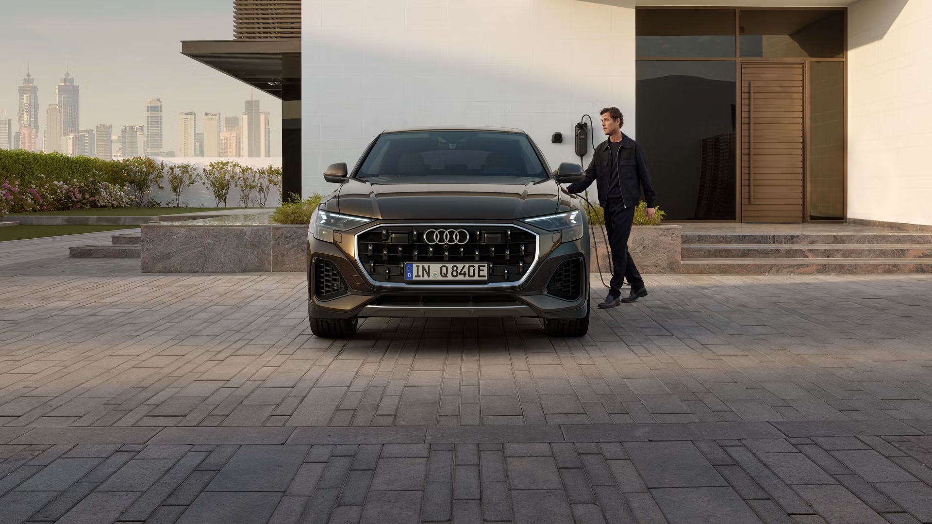 Frontansicht des Q8 SUV e-hybrid, dahinter ein modernes Gebäude. Neben den Fahrzeug steht ein modisch gekleideter junger Mann und hält den Stecker einer am Gebäude angebrachten Ladesäule in der Hand.