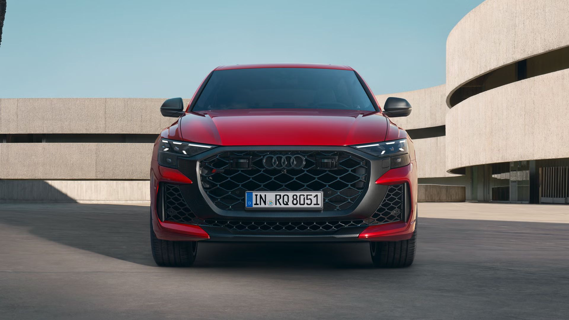 Der RS Q8 SUV performance in Chilirot Metallic von vorne parkend vor einem futuristischen Gebäude.