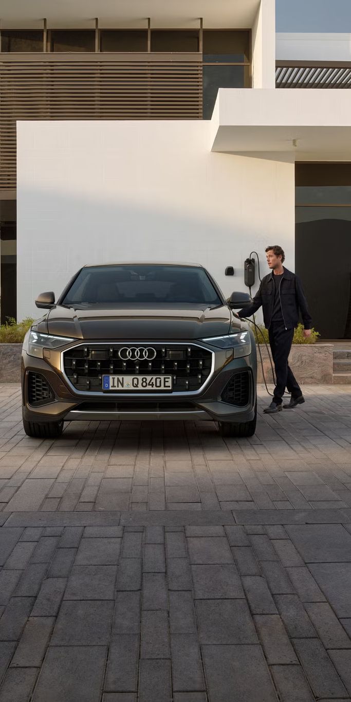 Frontansicht des Q8 SUV e-hybrid, dahinter ein modernes Gebäude. Neben den Fahrzeug steht ein modisch gekleideter junger Mann und hält den Stecker einer am Gebäude angebrachten Ladesäule in der Hand.