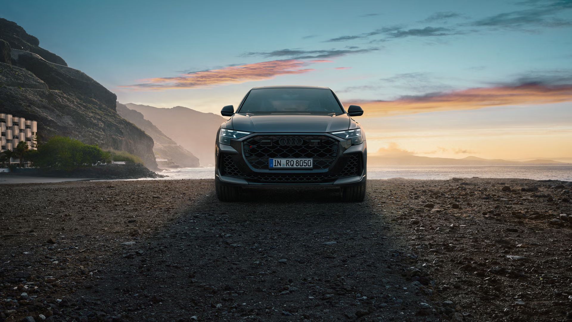 Der RS Q8 SUV von vorn an einem Strand auf Schotter, im Hintergrund das Meer und seitlich hervorragende Berge.
