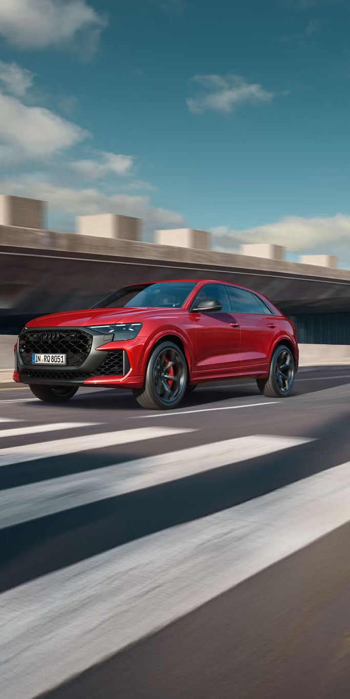 Der RS Q8 SUV performance schräg von vorn nähert sich einem Zebrastreifen. Rechts daneben ein futuristisches Gebäude in Betonoptik.