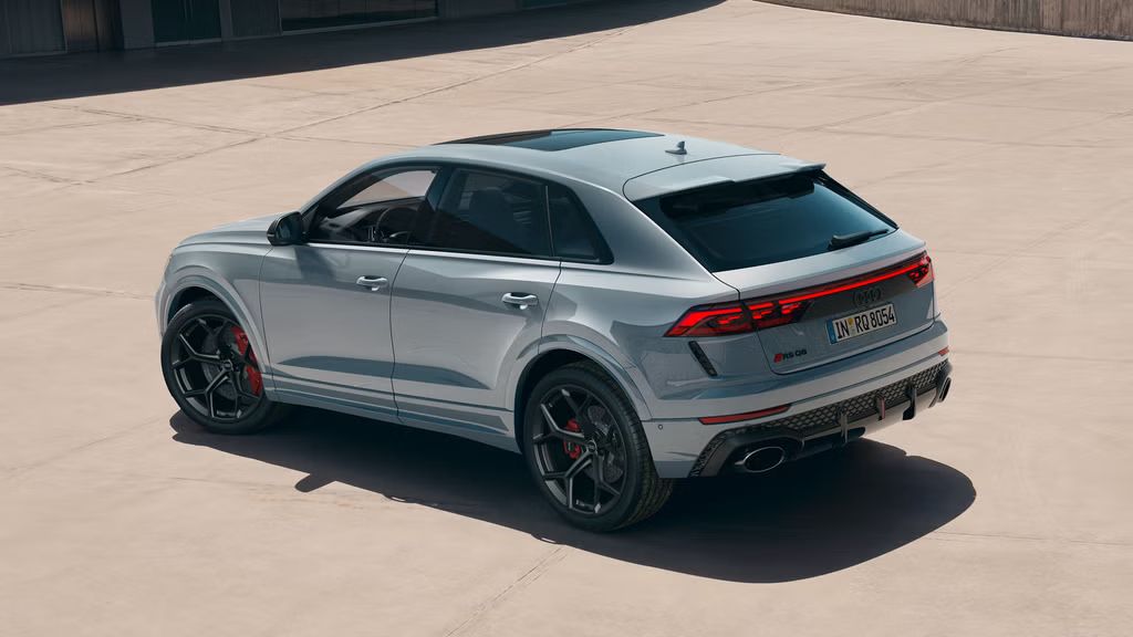 Der RS Q8 SUV performance mit Individuallackierung Audi exclusive in Pfeilgrau Perleffekt von schräg hinten auf einem sonnendurchfluteten Parkplatz vor einem modernen Gebäude.