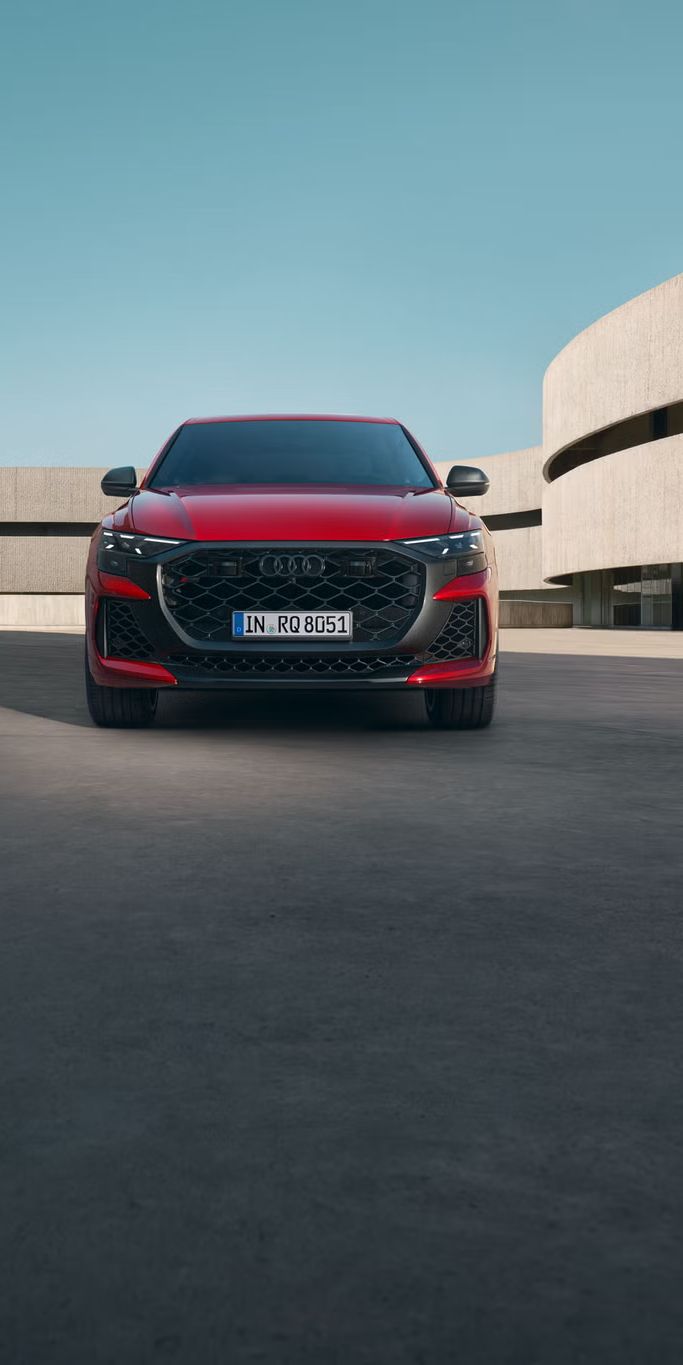 Der RS Q8 SUV performance in Chilirot Metallic von vorne parkend vor einem futuristischen Gebäude.