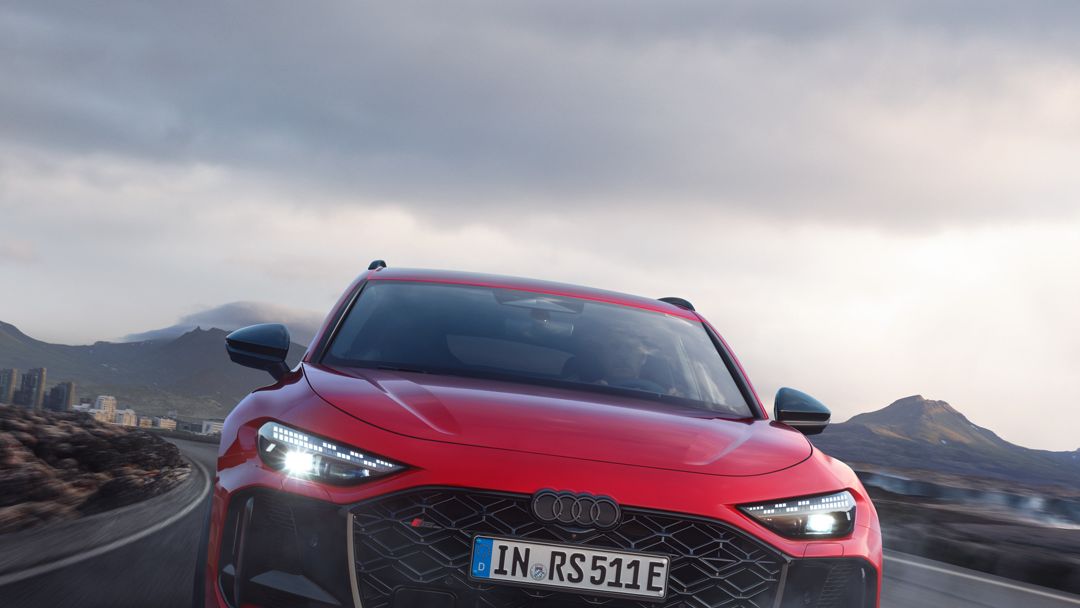 Un Audi RS 5 Avant roșu circulă pe un drum într-un peisaj muntos. Cerul e înnorat.