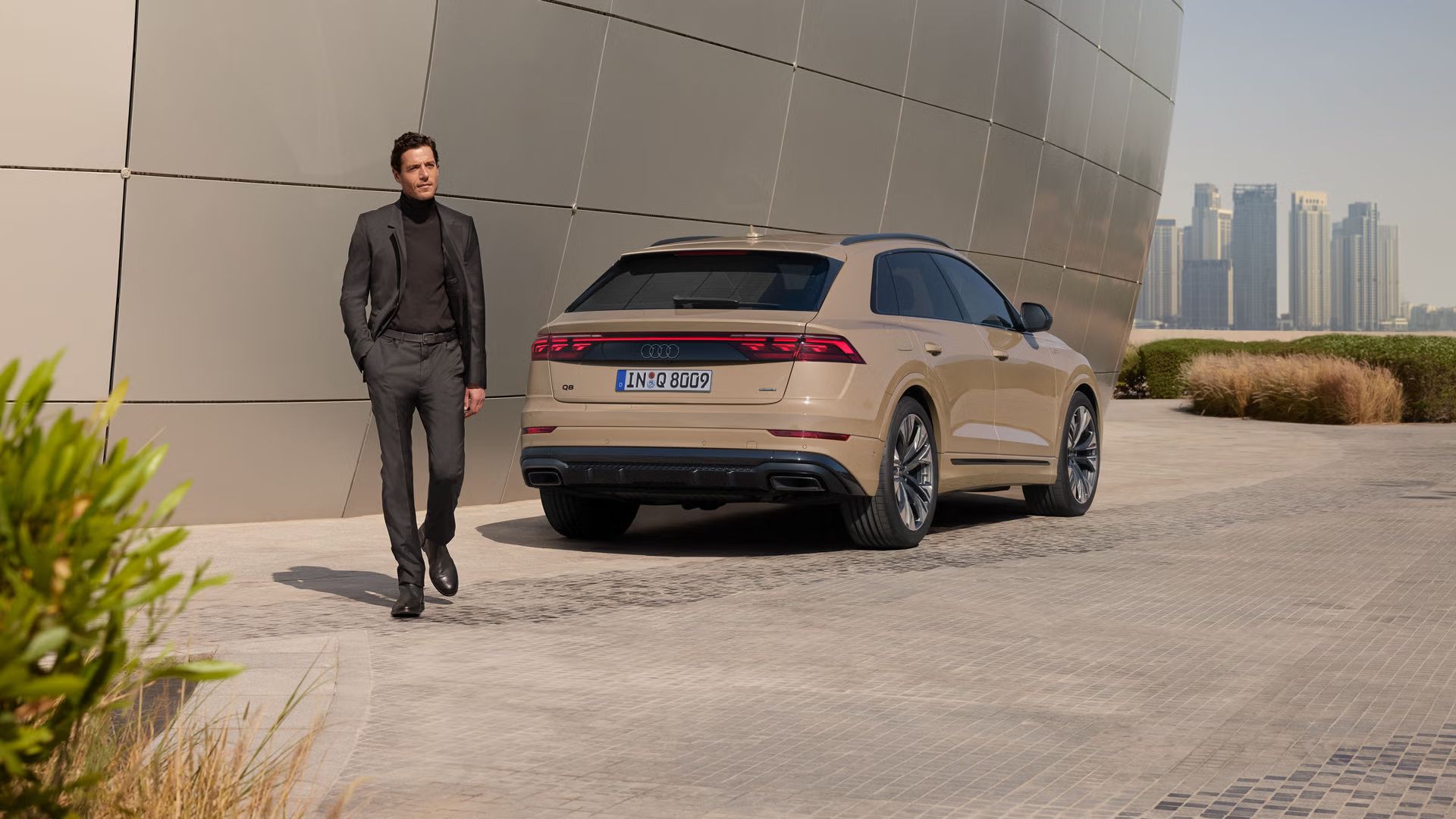 Der Q8 SUV schräg von hinten parkt an einem modernen Gebäude, ein modisch gekleideter junger Mann läuft vor dem Fahrzeug. Im Hintergrund die Skyline einer Großstadt.