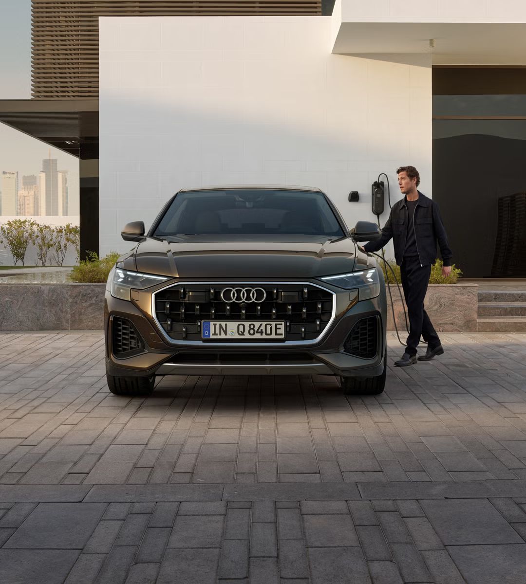 Frontansicht des Q8 SUV e-hybrid, dahinter ein modernes Gebäude. Neben den Fahrzeug steht ein modisch gekleideter junger Mann und hält den Stecker einer am Gebäude angebrachten Ladesäule in der Hand.