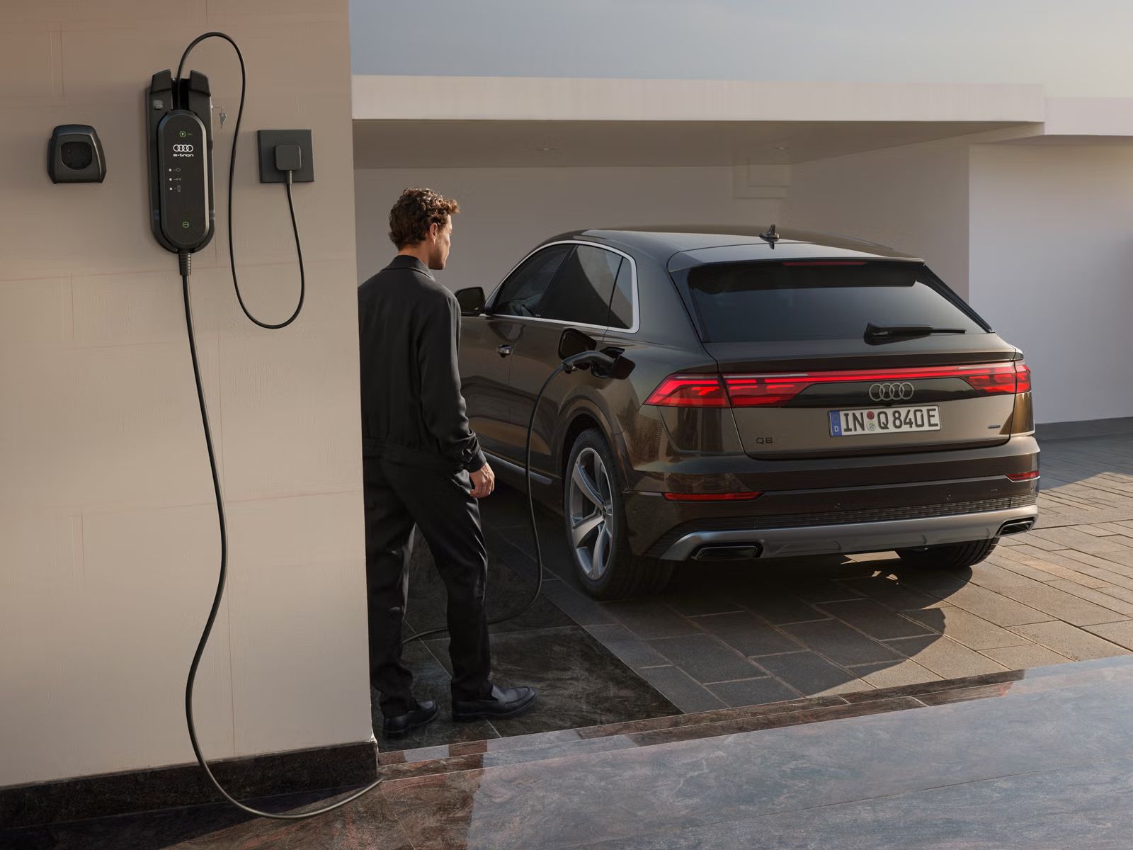 Der Q8 SUV e-hybrid schräg von hinten parkt vor einer weißen Mauer, im Vordergrund ein Gebäude, an dem eine Ladesäule angebracht ist. Ein modisch gekleideter junger Mann steht neben dem Fahrzeug.