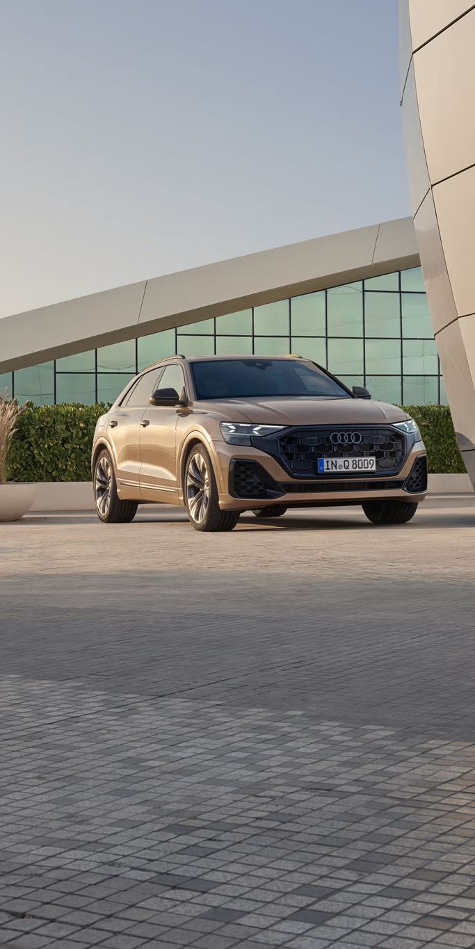 Der Q8 SUV schräg von vorn parkt vor einem modernen Gebäude. Eine bepflanzte Hecke trennt das Fahrzeug von dem dahinter liegenden, voll verglasten Gebäude.