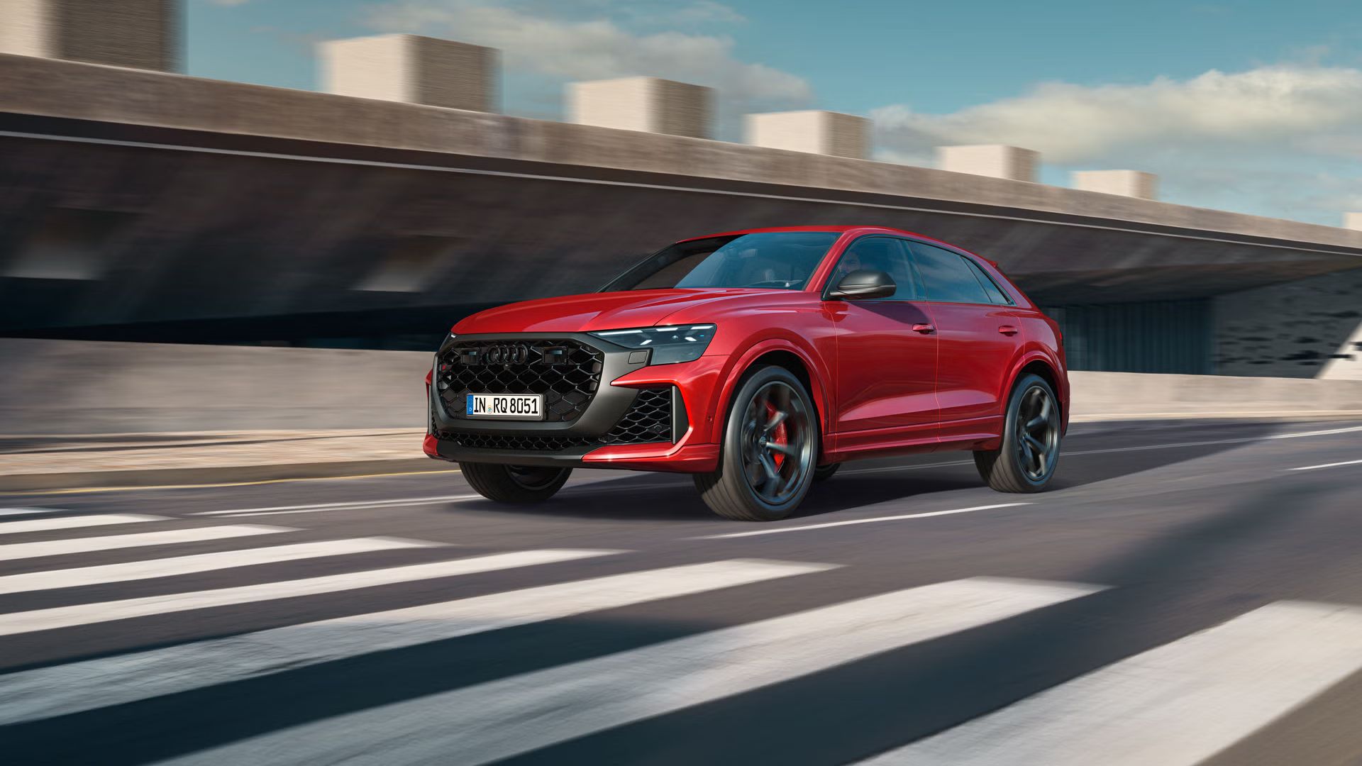 Der RS Q8 SUV performance schräg von vorn nähert sich einem Zebrastreifen. Rechts daneben ein futuristisches Gebäude in Betonoptik.