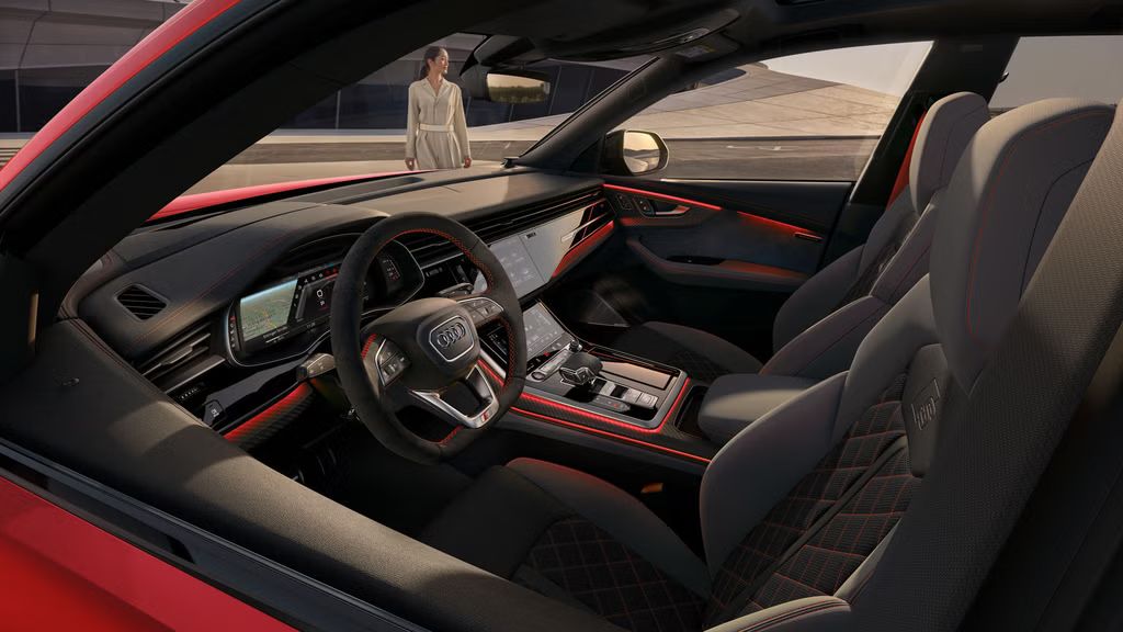 Blick in das Cockpit des SQ8 SUV mit dem Designpaket schwarz-neodymgold von der Fahrerseite, im Hintergrund ein futuristisches Gebäude mit geschwungener Form. Vor dem Fahrzeug steht eine modisch gekleidete junge Frau.